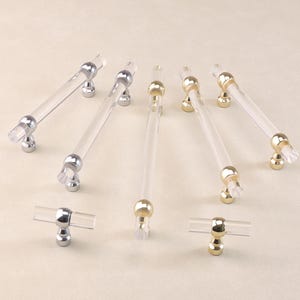 Könnte beinhalten: Sortiment an klaren Acryl-Schrankgriffen mit goldenen und silbernen Oberflächen. Die Griffe sind in verschiedenen Ausführungen, darunter Stangen- und T-Form.