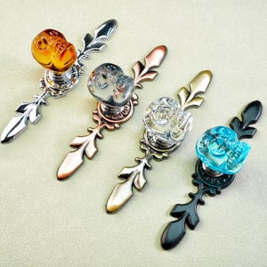 Op de afbeelding: Vier decoratieve kastknoppen met sierlijke metalen bases en kristallen of glazen knoppen. De knoppen zijn amber, helder en blauw. De metalen bases zijn zilver, koper, goud en zwart.