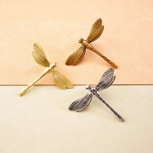 May include: Three dragonfly brooches in gold, bronze, and silver. The brooches feature detailed wing patterns and slender bodies. The background is a two-tone surface of beige and light green.
