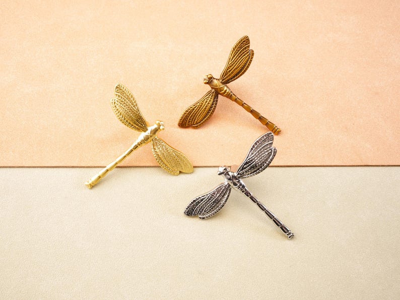 May include: Three dragonfly brooches in gold, bronze, and silver. The brooches feature detailed wing patterns and slender bodies. The background is a two-tone surface of beige and light green.