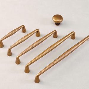 May include: A set of bronze-colored cabinet hardware, including four elongated pulls and one round knob. The hardware has a textured finish and a warm, metallic hue, suitable for kitchen or furniture upgrades.