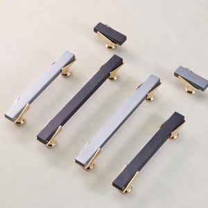 May include: A collection of modern cabinet hardware, including long bar pulls and smaller knobs. The pulls and knobs feature a rectangular design with a contrasting color and gold-toned accents. The hardware is designed for furniture and cabinetry.