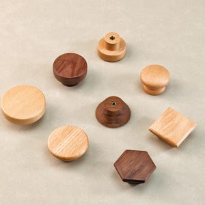 Może przedstawiać: Kolekcja drewnianych gałek do szafek w r&oacute;żnych kształtach i odcieniach drewna. Gałki obejmują okrągłe, kwadratowe i sześciokątne wzory, z jasnym i ciemnym wykończeniem drewna. Gałki są ułożone na neutralnym tle.