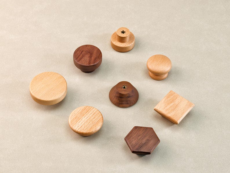 Może przedstawiać: Kolekcja drewnianych gałek do szafek w r&oacute;żnych kształtach i odcieniach drewna. Gałki obejmują okrągłe, kwadratowe i sześciokątne wzory, z jasnym i ciemnym wykończeniem drewna. Gałki są ułożone na neutralnym tle.