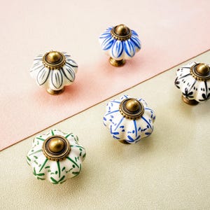 May include: Five ceramic cabinet knobs with floral patterns in white, blue, green, and black. Each knob has a brass-coloured base and top. The knobs are arranged on a two-tone pink and beige surface.