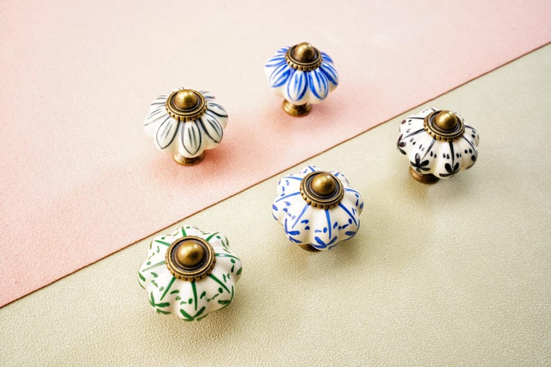 May include: Five ceramic cabinet knobs with floral patterns in white, blue, green, and black. Each knob has a brass-coloured base and top. The knobs are arranged on a two-tone pink and beige surface.