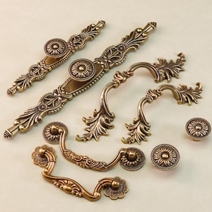 May include: An assortment of antique brass-colored cabinet hardware, including ornate pulls and knobs. The hardware features floral and scrollwork designs. The image shows various sizes and styles of the decorative hardware.