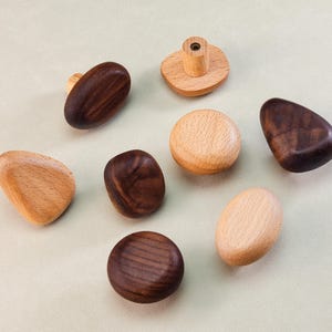 Op de afbeelding: Een assortiment houten kastknoppen in verschillende vormen en afwerkingen. Sommige zijn licht van kleur, andere zijn donkerbruin. De knoppen zijn gerangschikt op een lichtgroen oppervlak.