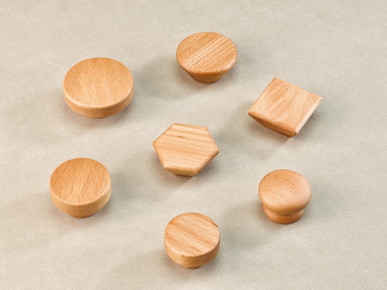 Może przedstawiać: Zestaw siedmiu drewnianych gałek do szafek w r&oacute;żnych kształtach, w tym okrągłych, sześciokątnych i kwadratowych. Gałki wykonane są z jasnego drewna i ułożone na neutralnym tle.
