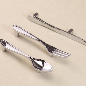 May include: A set of three silver cabinet pulls, each shaped like cutlery: a spoon, fork, and knife. The pulls have a polished finish and are designed to resemble silverware. The handles are mounted on short, cylindrical posts.