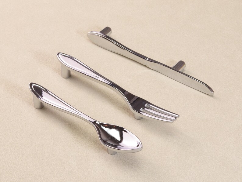 May include: A set of three silver cabinet pulls, each shaped like cutlery: a spoon, fork, and knife. The pulls have a polished finish and are designed to resemble silverware. The handles are mounted on short, cylindrical posts.