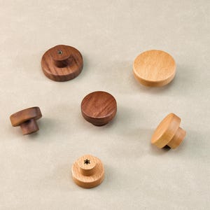 Może przedstawiać: Kolekcja drewnianych gałek do szafek w r&oacute;żnych kształtach i odcieniach drewna. Gałki mają wykończenie z ciemnego orzecha włoskiego, średniego brązu i jasnego klonu. Niekt&oacute;re gałki mają płaską g&oacute;rę, inne zaokrąglony lub grzybkowy kształt.