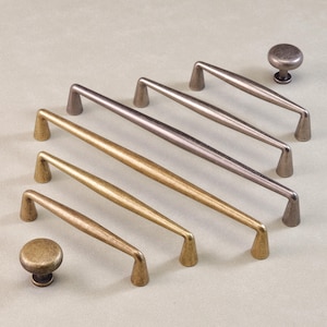 Könnte beinhalten: Eine Auswahl an Schrankbeschlägen, darunter knauf- und griffartige Elemente in Bronzetönen. Die Griffe variieren in der Größe und haben ein konisches Design. Die Hardware ist diagonal auf einer hellfarbigen Oberfläche angeordnet.