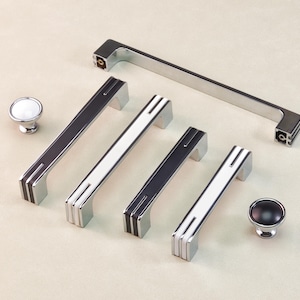 May include: A collection of cabinet hardware, including silver and black or white drawer pulls and knobs. The pulls have a rectangular design with a central stripe and are arranged with two knobs.