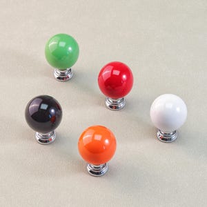 Peut inclure: Cinq boutons de meuble color&eacute;s. Les boutons sont ronds avec une finition brillante, en vert, rouge, noir, orange et blanc. Chaque bouton est mont&eacute; sur une base argent&eacute;e. Les boutons sont probablement destin&eacute;s aux meubles ou &agrave; la d&eacute;coration.