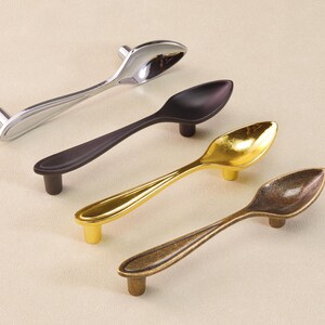 May include: Four spoon-shaped cabinet pulls in silver, black, gold, and bronze. Each pull has a spoon bowl design with a small, cylindrical base for mounting. The pulls are arranged diagonally, showing their different finishes.