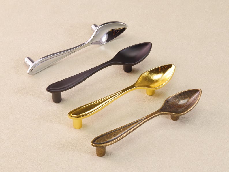 May include: Four spoon-shaped cabinet pulls in silver, black, gold, and bronze. Each pull has a spoon bowl design with a small, cylindrical base for mounting. The pulls are arranged diagonally, showing their different finishes.