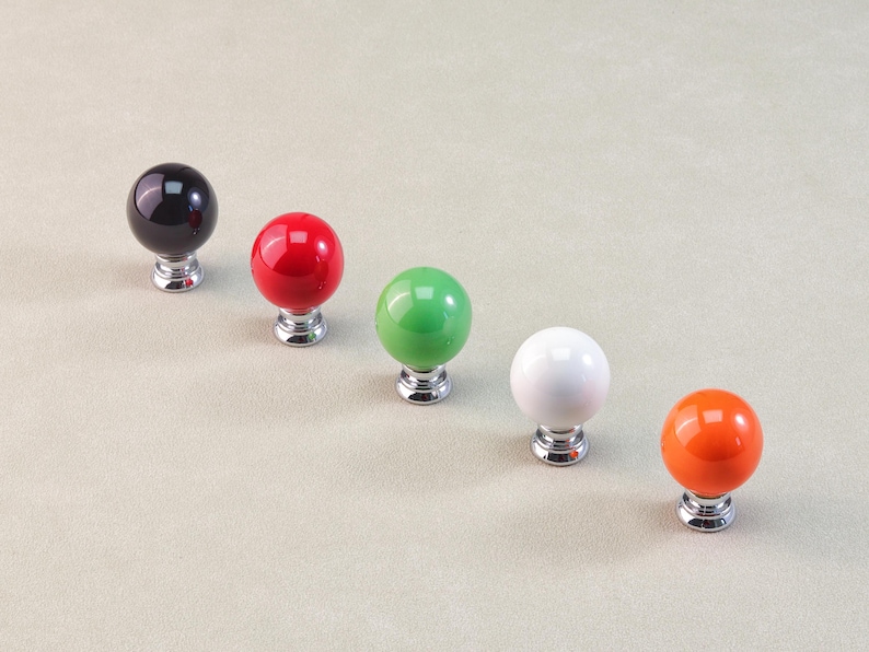 Peut inclure: Cinq boutons de meuble color&eacute;s en rang&eacute;e. Les boutons sont ronds avec une finition brillante, en noir, rouge, vert, blanc et orange. Chaque bouton est mont&eacute; sur une base argent&eacute;e.
