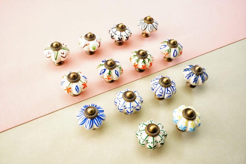 May include: A selection of ceramic cabinet knobs with floral patterns. The knobs have a white base with blue, orange, and green designs, and a brass-coloured metal cap. They are arranged on a pink and beige surface. Each knob is approximately 4 cm in diameter.