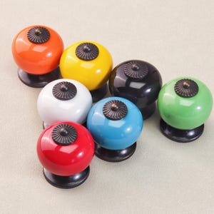 May include: A selection of seven brightly coloured cabinet knobs. Each knob is a different colour: orange, yellow, white, black, blue, green, and red. The knobs have a round shape with a decorative, dark metal top and a black base.