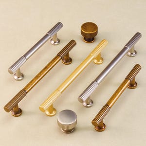 May include: A selection of cabinet handles and knobs in various metallic finishes, including silver, gold, and bronze. The handles feature a ribbed design, and the knobs are circular with a matching texture. The hardware is displayed on a neutral surface.