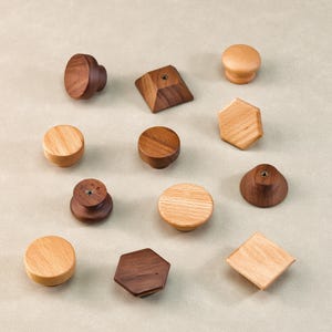 May include: A selection of wooden cabinet knobs in diverse shapes and wood tones. The knobs feature round, geometric, and mushroom-shaped designs, displaying natural wood grain and various finishes. The knobs are arranged on a neutral background.
