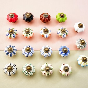 May include: A selection of decorative ceramic cabinet knobs in various colours and patterns. The knobs are flower-shaped with brass hardware. Colours include red, black, brown, green, white, blue, and cream. The knobs are arranged on a pink and beige background.