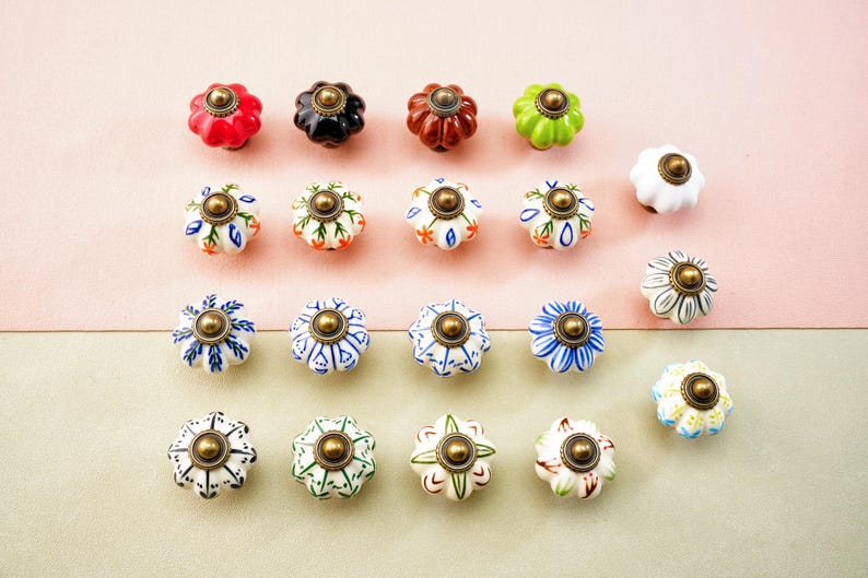 May include: A selection of decorative ceramic cabinet knobs in various colours and patterns. The knobs are flower-shaped with brass hardware. Colours include red, black, brown, green, white, blue, and cream. The knobs are arranged on a pink and beige background.