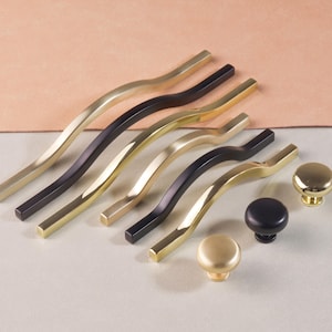 May include: A collection of gold and black cabinet hardware, including curved bar pulls and round knobs. The hardware is displayed on a light surface with a peach-colored background, showcasing various design options.