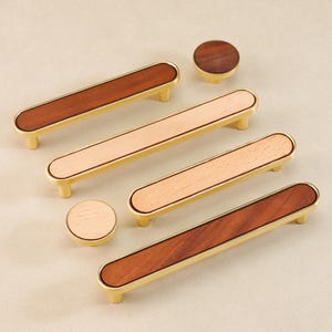 Op de afbeelding: Een verzameling kastbeslag, waaronder rechthoekige handgrepen en ronde knoppen. De handgrepen hebben een licht houten midden met een donker of licht houten accent, gezet in een goudkleurig frame. De knoppen hebben een bijpassend ontwerp.