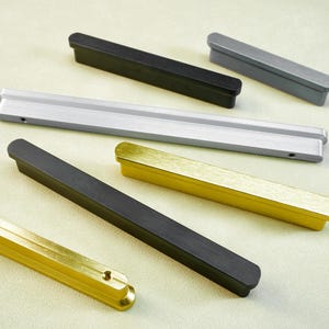 May include: A selection of contemporary cabinet pulls in different colours. The pulls are rectangular with rounded ends, in black, silver, and gold tones. The hardware is designed for drawers and cabinets.