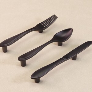 May include: Three dark bronze-coloured cabinet pulls shaped like a fork, spoon, and knife. The pulls have a simple, elegant design with a rounded handle and a short, cylindrical base for mounting. Ideal for kitchen or dining room furniture.