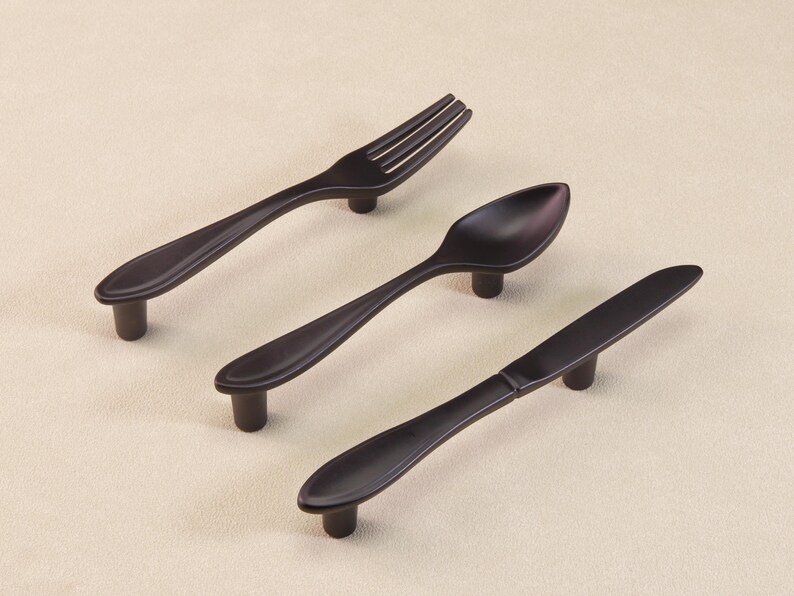 May include: Three dark bronze-coloured cabinet pulls shaped like a fork, spoon, and knife. The pulls have a simple, elegant design with a rounded handle and a short, cylindrical base for mounting. Ideal for kitchen or dining room furniture.