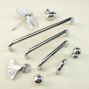 May include: A collection of shiny silver cabinet hardware, including various handle styles, knobs, and a decorative dragonfly. The hardware is arranged on a light beige surface, showcasing the reflective quality of the metal.