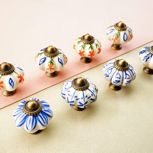 May include: Eight ceramic cabinet knobs with floral designs. The knobs have a white base with patterns in blue, orange, and green. Each knob has a bronze-coloured metal base and top. The knobs are displayed on a two-tone pink and beige surface.