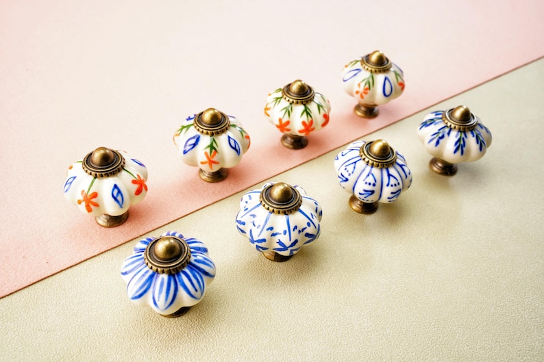 May include: Eight ceramic cabinet knobs with floral designs. The knobs have a white base with patterns in blue, orange, and green. Each knob has a bronze-coloured metal base and top. The knobs are displayed on a two-tone pink and beige surface.