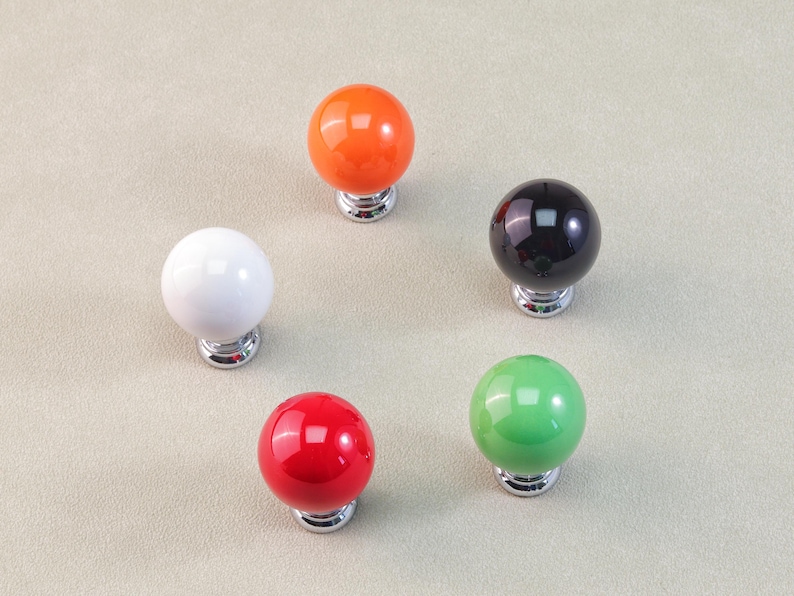 Peut inclure: Cinq boutons de meuble color&eacute;s dispos&eacute;s en cercle. Les boutons sont brillants et sph&eacute;riques, avec des couleurs comprenant le blanc, l'orange, le noir, le rouge et le vert. Chaque bouton a une base argent&eacute;e.