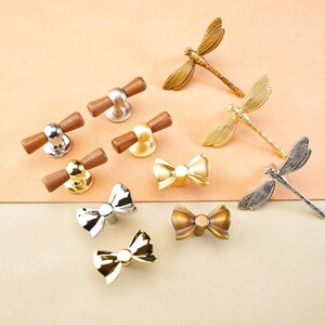 May include: A selection of decorative cabinet hardware, including knobs and dragonfly and bow-shaped pulls. The knobs have wood and metal finishes in silver, gold, and brass. The dragonfly and bow pulls are in gold, silver, and bronze tones.