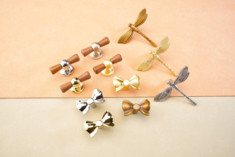 May include: A selection of decorative cabinet hardware, including knobs and dragonfly and bow-shaped pulls. The knobs have wood and metal finishes in silver, gold, and brass. The dragonfly and bow pulls are in gold, silver, and bronze tones.