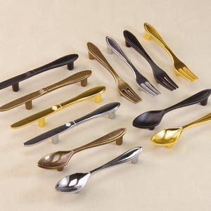 May include: A collection of cabinet handles designed to resemble cutlery. The selection includes handles shaped like spoons, forks, and knives in various metallic finishes, including gold, silver, and black. Suitable for kitchen or dining room furniture.