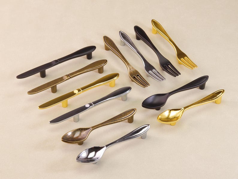 May include: A collection of cabinet handles designed to resemble cutlery. The selection includes handles shaped like spoons, forks, and knives in various metallic finishes, including gold, silver, and black. Suitable for kitchen or dining room furniture.