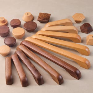 Könnte beinhalten: Sortiment an Holzknauf- und Schubladengriffen in verschiedenen Formen und Größen. Die Knöpfe sind rund, quadratisch und sechseckig, während die Griffe lang und gebogen sind. Das Holz ist hellbraun.