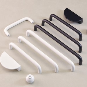 May include: A selection of contemporary cabinet hardware in white and black. The set includes curved bar pulls, half-moon pulls, and a round knob. The hardware is made of metal and is shown on a neutral background.