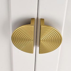 Könnte beinhalten: Zwei goldfarbene Schranktürgriffe mit konzentrischem Kreisdesign. Die Griffe sind Halbkreise und auf einer weißen Oberfläche montiert.