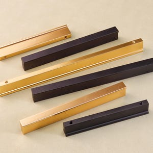 Puede incluir: Una variedad de tiradores rectangulares para armarios en dorado y negro. Los tiradores están dispuestos diagonalmente, mostrando sus diferentes longitudes y acabados. Cada tirador tiene un diseño elegante y moderno, con agujeros pretaladrados.