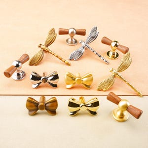 May include: A selection of decorative cabinet hardware, featuring dragonfly and bow-tie designs. The knobs and pulls are made of metal in gold, silver, and bronze, with wooden elements. The items are displayed on a light peach surface.