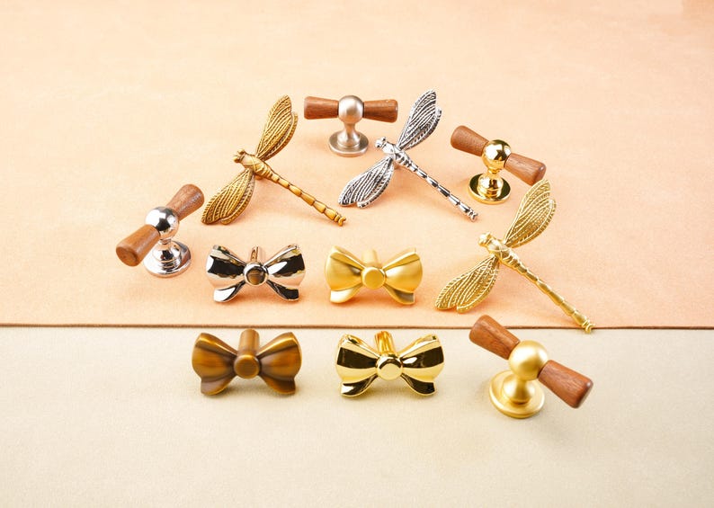 May include: A selection of decorative cabinet hardware, featuring dragonfly and bow-tie designs. The knobs and pulls are made of metal in gold, silver, and bronze, with wooden elements. The items are displayed on a light peach surface.