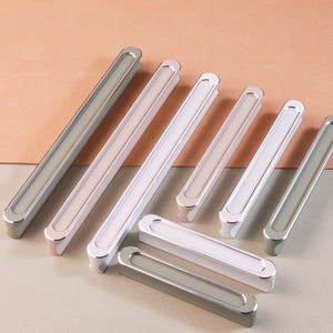 May include: A set of six cabinet pulls in silver and white finishes. The pulls are rectangular with rounded edges and a recessed center.
