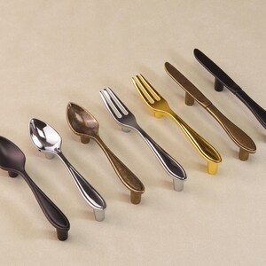 May include: Cabinet hardware in spoon, fork, and knife designs. The handles are in various metallic finishes: black, silver, gold, and bronze. The items are arranged diagonally on a neutral background. The hardware is suitable for kitchen or dining room cabinets.