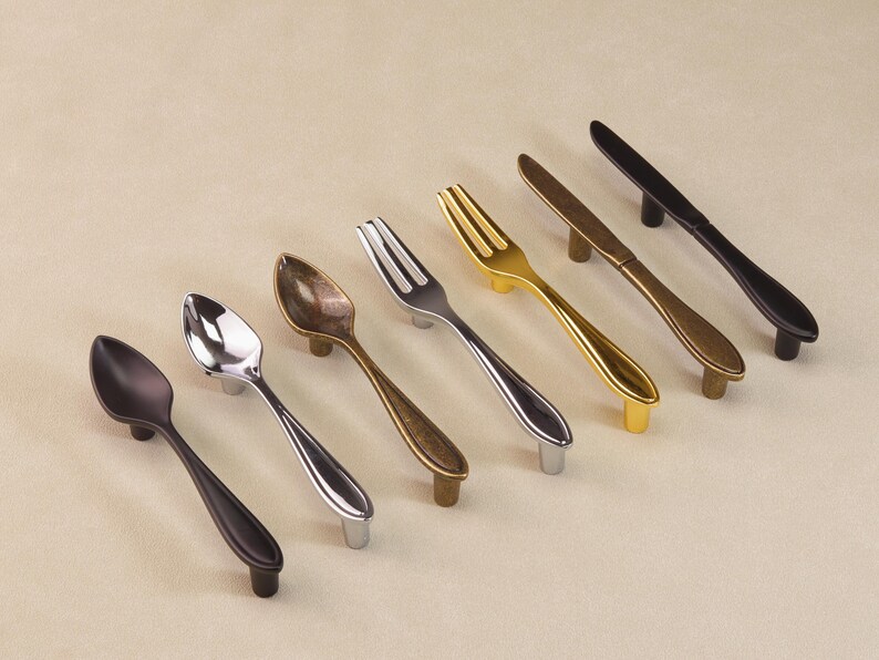 May include: Cabinet hardware in spoon, fork, and knife designs. The handles are in various metallic finishes: black, silver, gold, and bronze. The items are arranged diagonally on a neutral background. The hardware is suitable for kitchen or dining room cabinets.